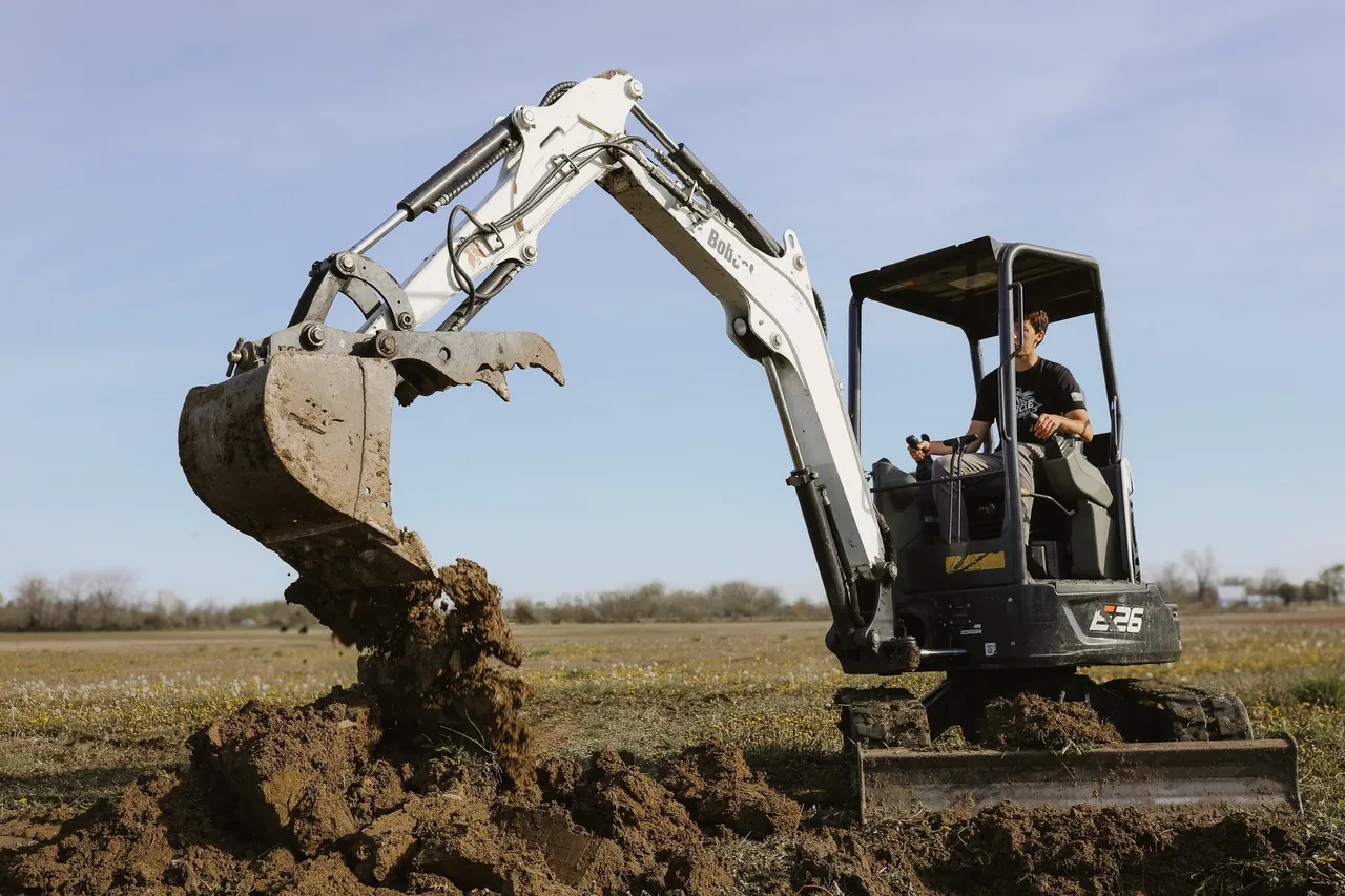 A mini excavator digging in an open field, dirt spilling from its bucket onto the pile below